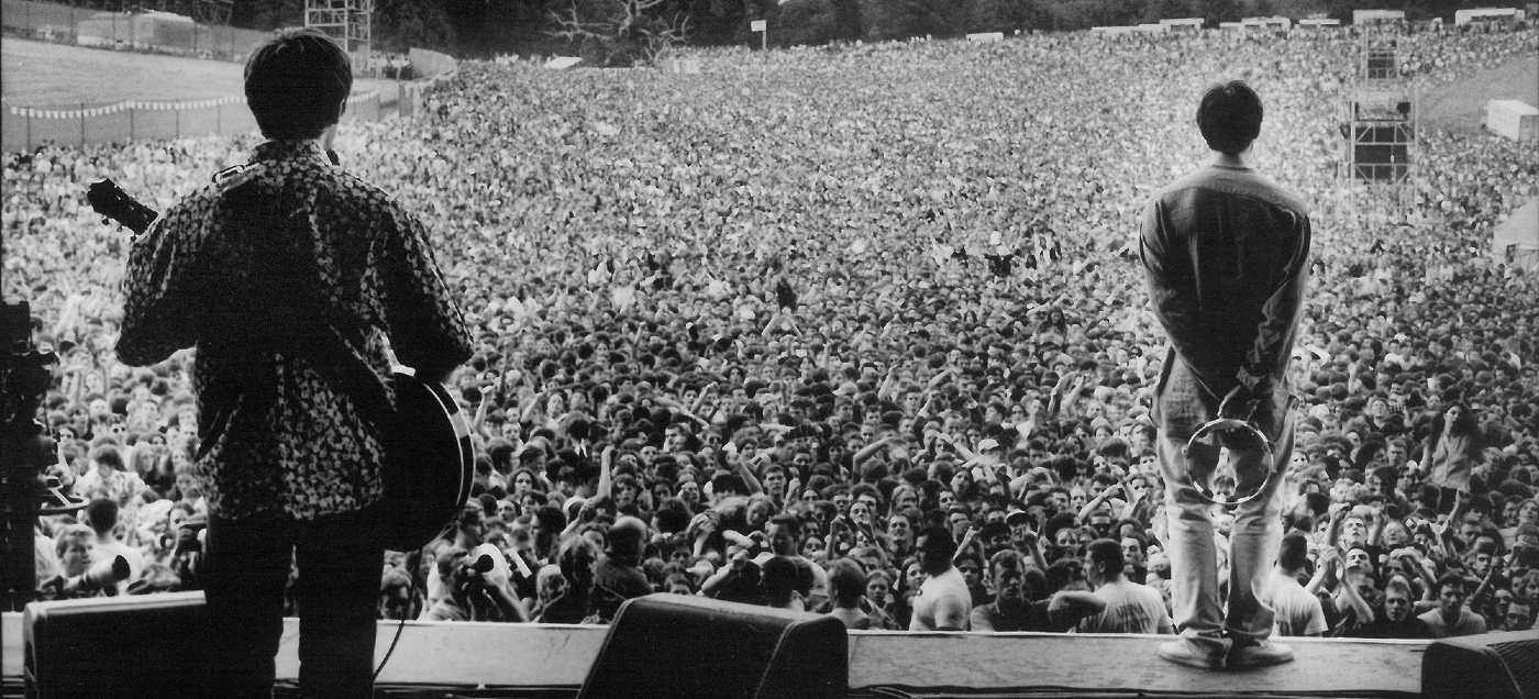 Black and white photo from behind Noel on the left and Liam on the right, both on stage, looking out over a sea of people