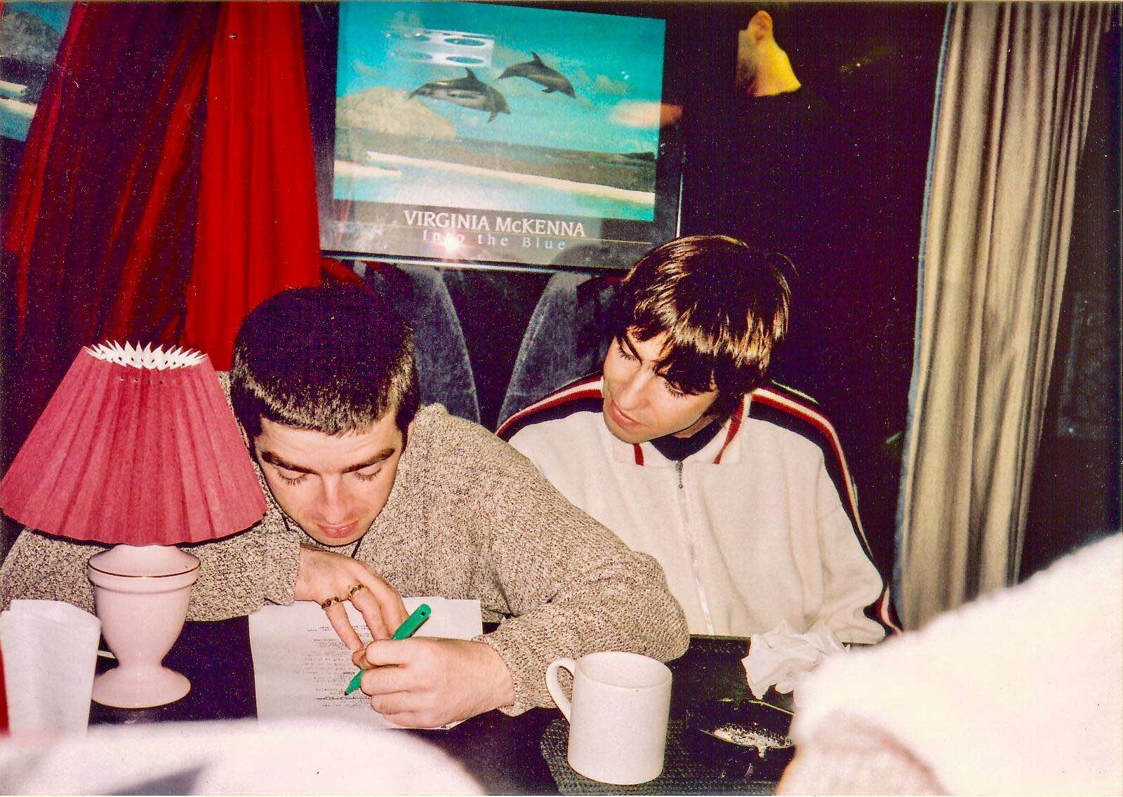 Noel at a table on a tour bus, writing on a sheet of paper with a green pen and a red lamp in frint of him, with Liam sitting close looking over his shoulder