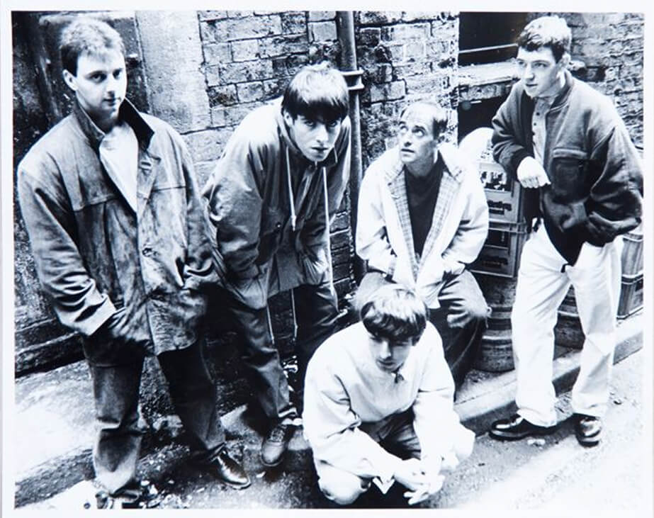 Black and white photo of the band in 1991, stading and crouched in what appears to be an alleyway, all looking in various directions with Liam being the only one looking into camera