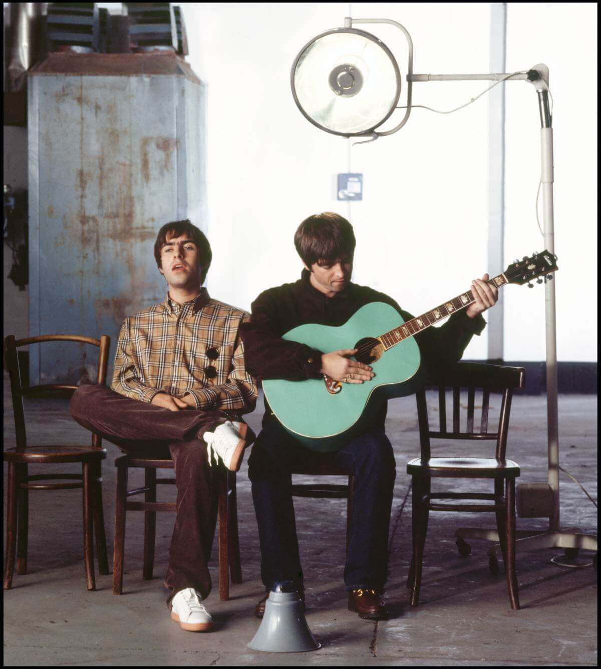 Liam and Noel sitting on 2 of 4 chairs in a row in a warehouse style setting, Noel in playing a guitar while Liam sings, a megaphone rests on the floor in front of them