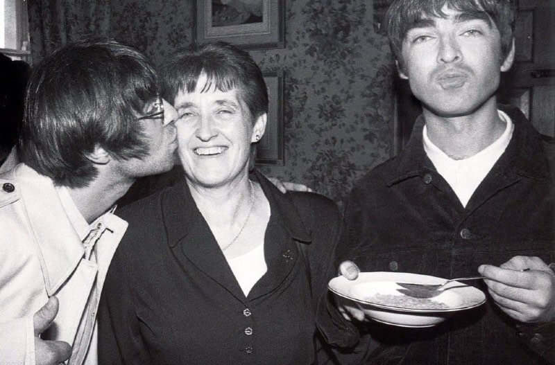 A black and white photo of Liam on the left, leaning in a kissing his smiling mum on the cheek, with Noal on the right eating a bowl of cereal and puckering his lips