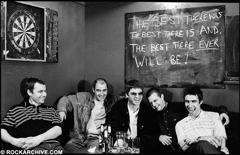 Black and white photo of the band all sitting at a table of beer and glasses, laughing, in the corner of a pub. Above them to the left is a dartboard and to the right a blackboard with the words 'The best there was the best there is and, the best there ever will be!