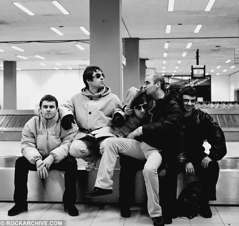 A black and white photo of the band all sitting on an empty bannge carousel at an airport, Liam is looking off the side, Noel has a cowboy hat on with Paul 'Bonehead' Arthurs sitting on his lap, kissing the hat